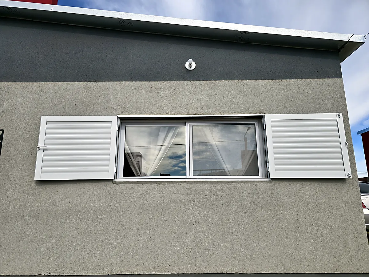 Ventana con postigos de aluminio blanco en una fachada gris